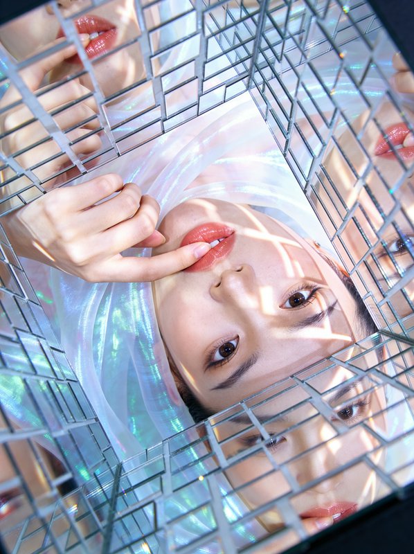 Young woman peering through a mirrored grid cube with iridescent lighting, her lips and hand visi...
