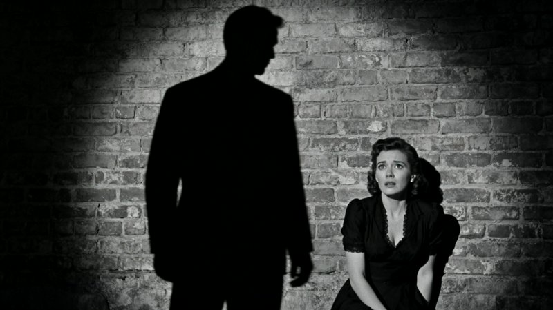 Black-and-white film-noir scene: a terrified woman sits by a brick wall as a shadowy man looms nearby.