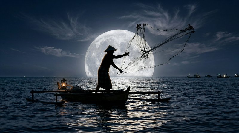 Silhouette of a fisherman in a small boat casting a net under a full moon over a moonlit sea.