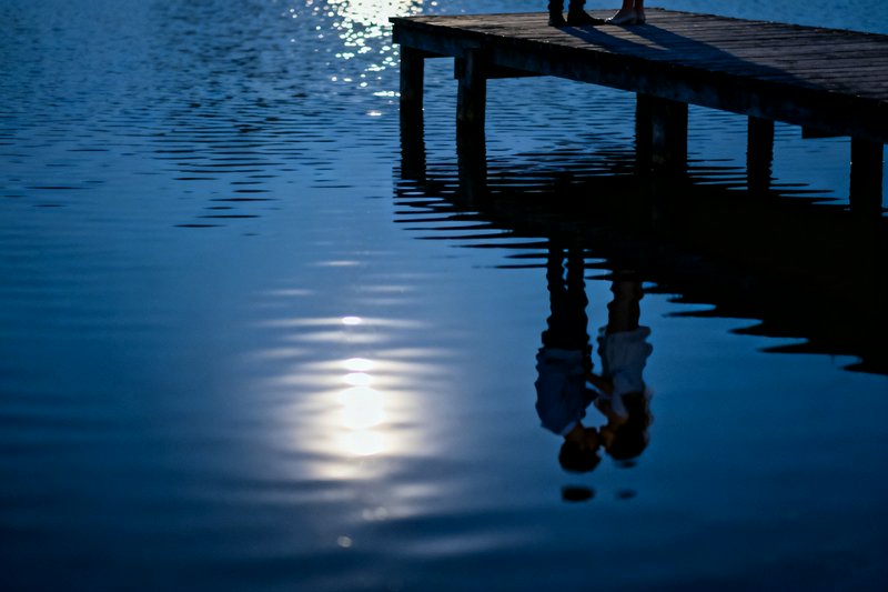 Silhouette of a couple standing on a wooden dock reflected in a calm blue lake at dusk.