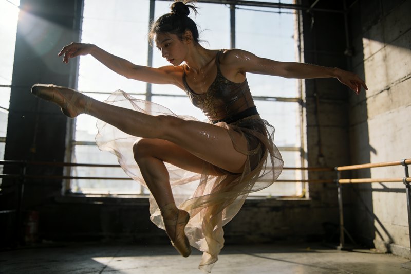 Female dancer mid-jump in an industrial studio, wearing a sheer skirt and leotard with sunlight s...