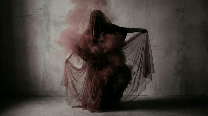 Silhouetted figure draped in red veil with smoky haze against a textured wall.