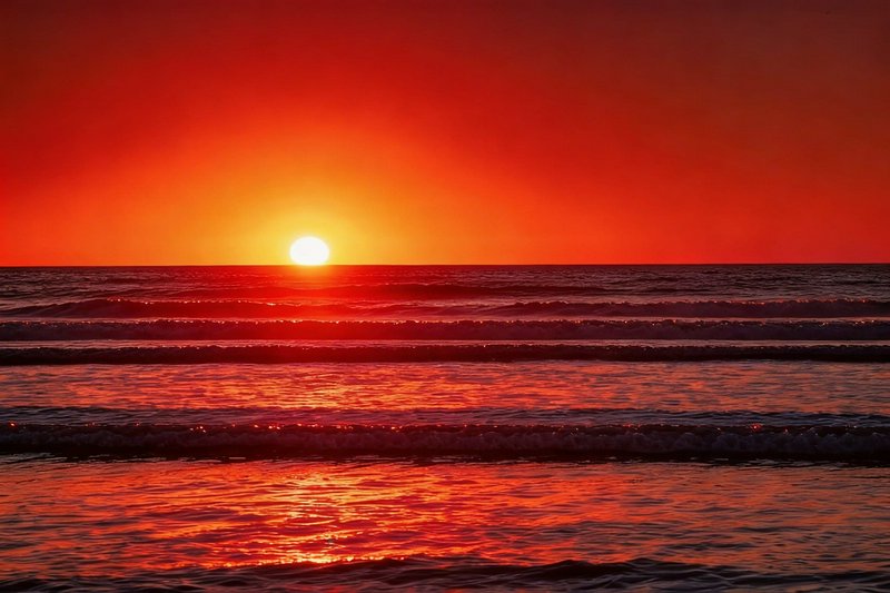 Sunset over the ocean with an orange-red sky and gentle waves on the shore