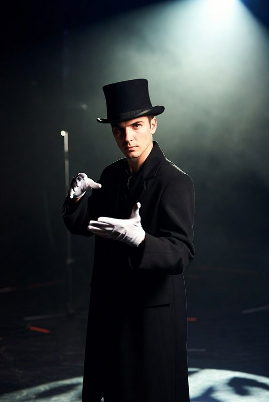 Magician in a black coat and top hat with white gloves performing on stage under dramatic spotlight.