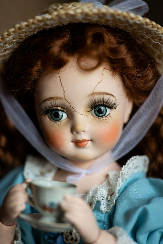 Close-up of vintage porcelain doll with blue eyes, cracked face, lace bonnet, holding a teacup.