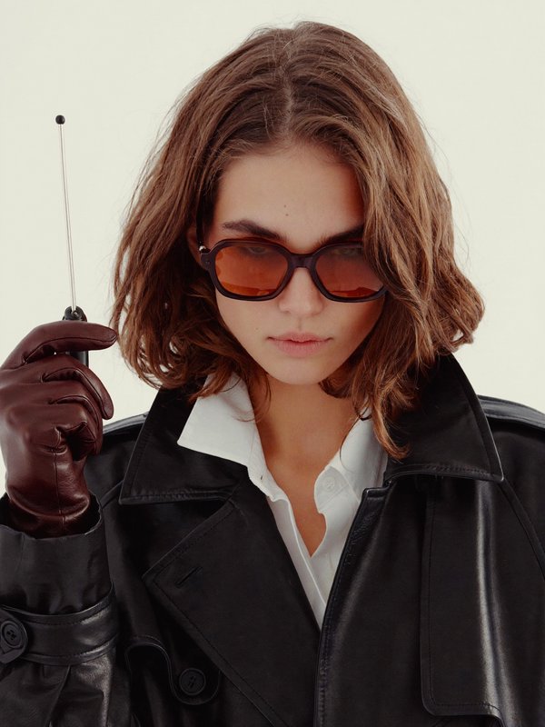 Woman in a black leather trench, brown gloves, and sunglasses holding a slim antenna.