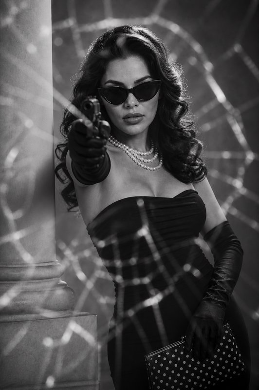 Stylish woman in a black gown, pearls, and cat-eye sunglasses points a pistol through shattered glass.