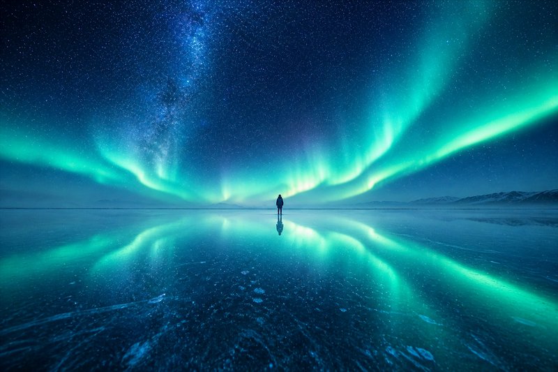 Silhouette of a person standing on a reflective icy plain under vibrant green and blue aurora bor...