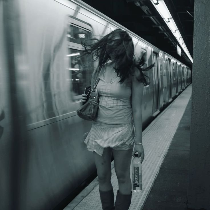 Woman on a subway platform with motion blur, standing beside a moving train while holding a water bottle and purse.