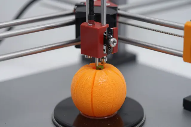 Close-up of a 3D printer extruding filament to build a textured spherical orange print on a circular build plate