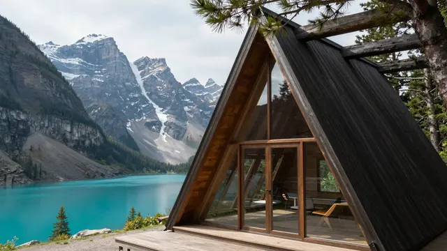 Modern A-frame cabin with floor-to-ceiling glass overlooks a turquoise alpine lake and snow-dusted peaks