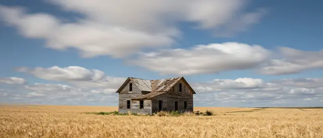 Weathered abandoned wooden farmhouse stands in a golden wheat field beneath streaking long exposure clouds