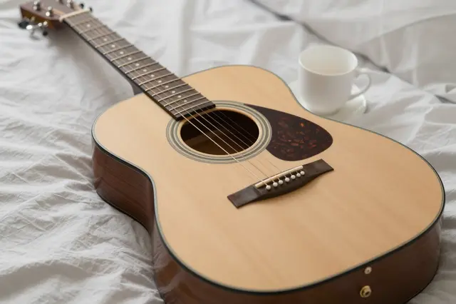 An acoustic guitar and a white cup of tea rest on rumpled soft white bed linens.