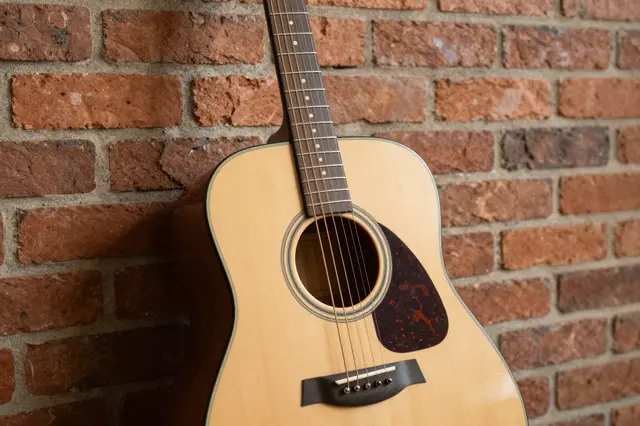 A natural wood acoustic guitar leans against a textured red brick wall under warm soft light.