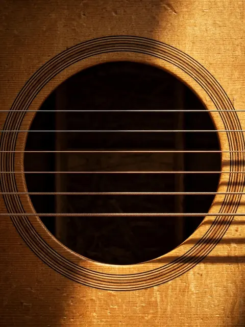 Close up of an acoustic guitar round sound hole with crossing strings lit by warm side light on golden wood