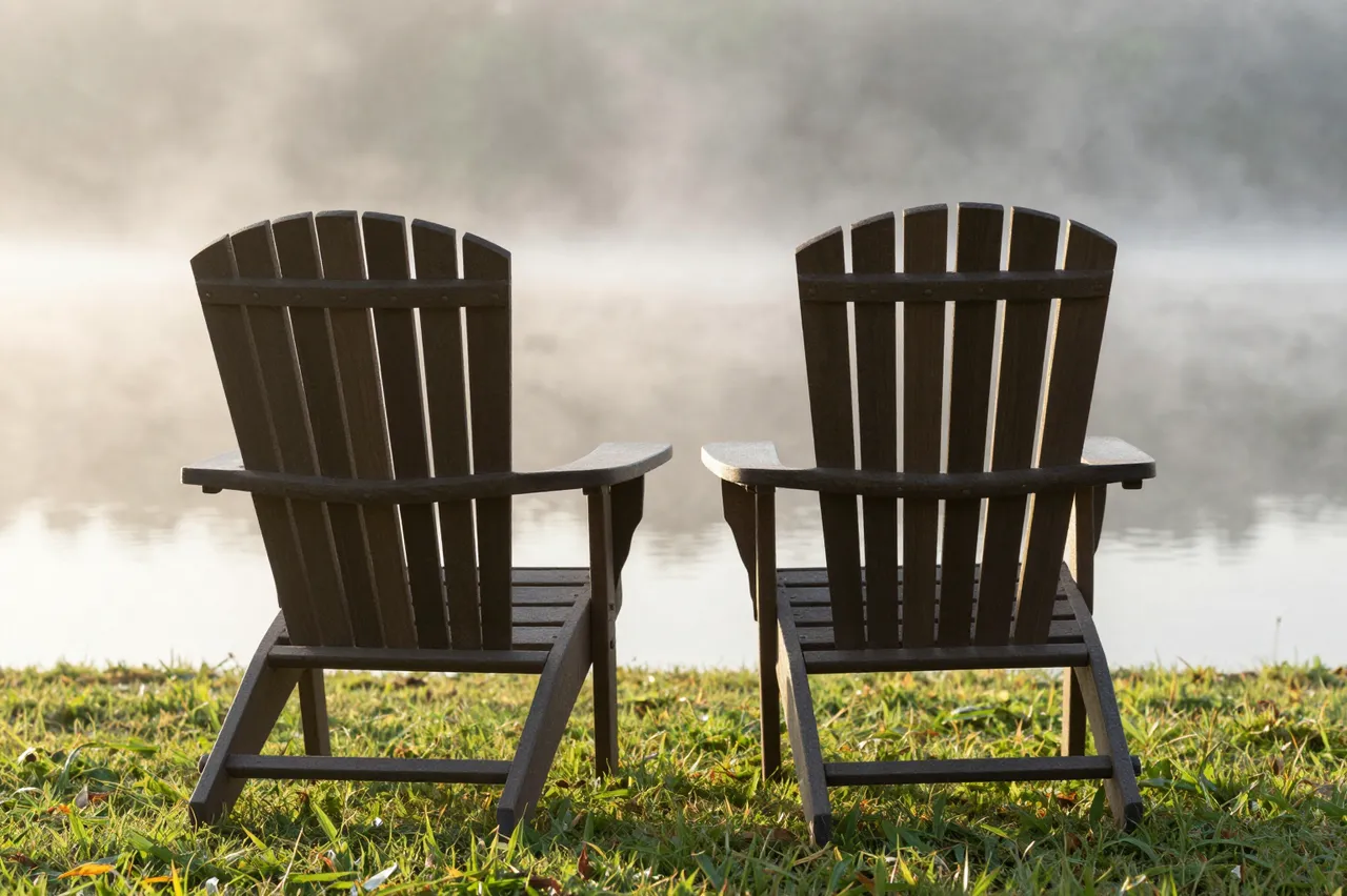 Two wooden lawn chairs sit side by side on grass facing a misty sunlit lake shore.