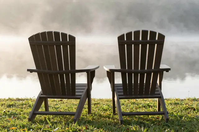 Two wooden lawn chairs sit side by side on grass facing a misty sunlit lake shore.