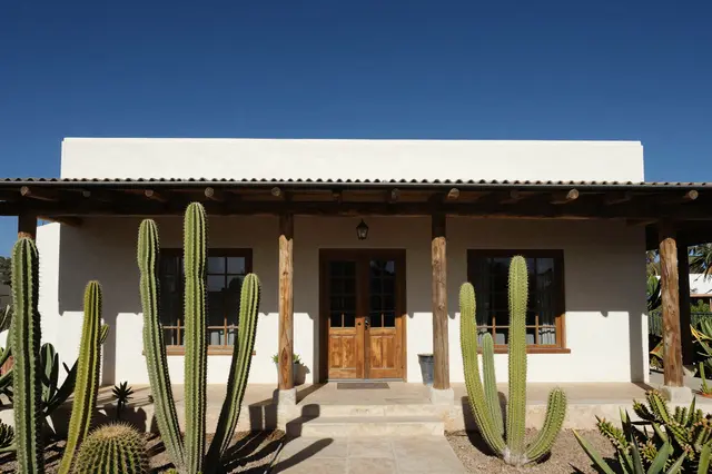 White adobe home with wooden porch beams and desert cacti stands under a deep clear blue sky.