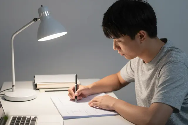 Young adult man writes in a notebook at a desk lit by a desk lamp at night