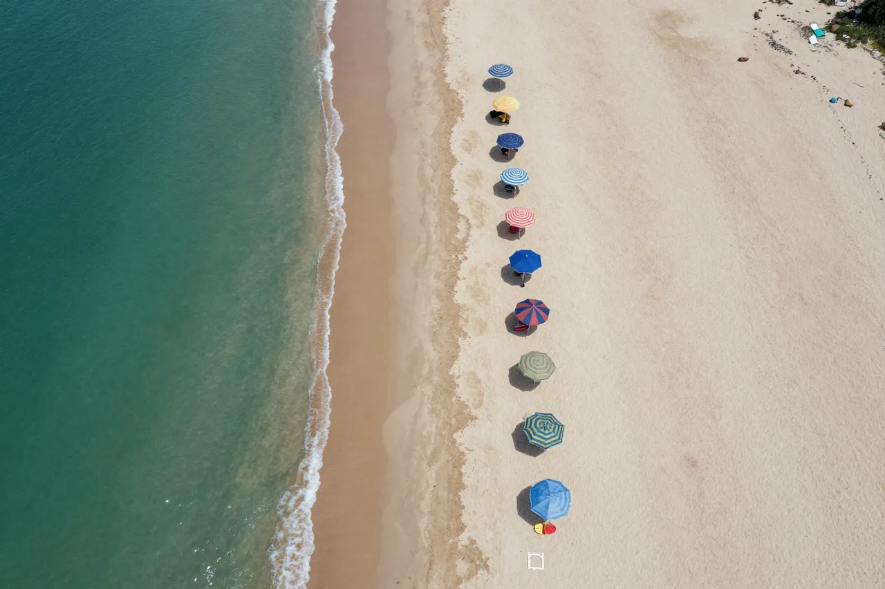 Top-down drone view of a sandy beach lined with bright umbrellas next to turquoise ocean water.