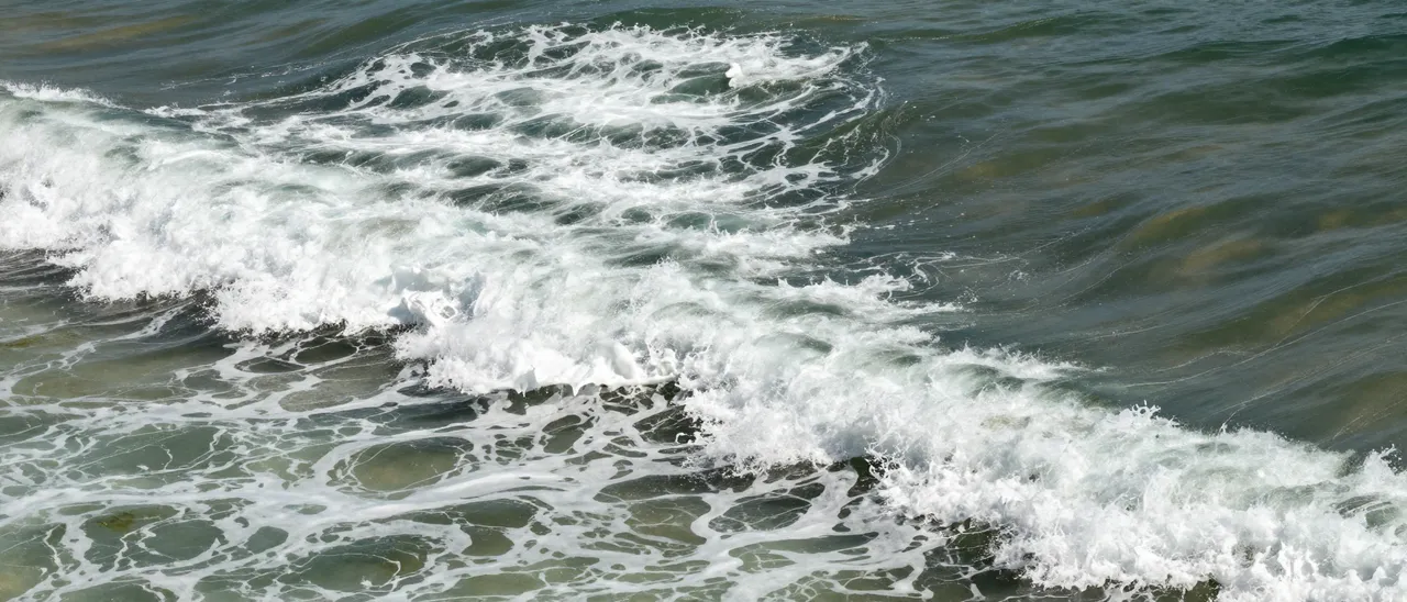 Aerial view of curling ocean waves forming bright white foamy surf across greenish coastal water.