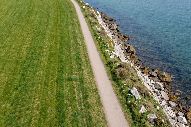 Aerial view of a paved footpath running between green grassy fields and a rocky sea cliff edge beside calm blue coastal water