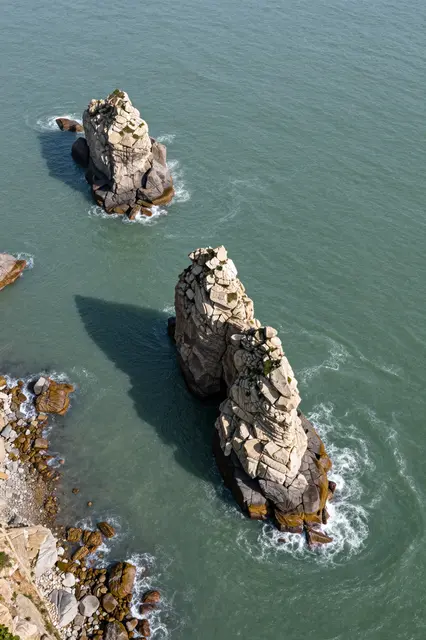 A high-angle aerial view of rugged sea stacks rising from calm green coastal water beside a rocky shoreline.