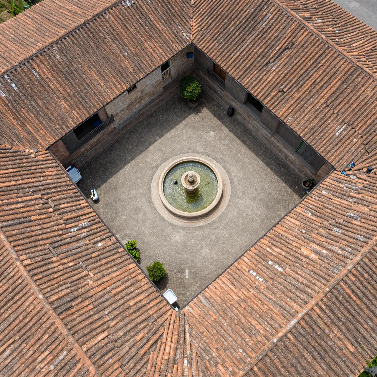 Top-down aerial view of a square courtyard with a central fountain surrounded by weathered terracotta rooftops