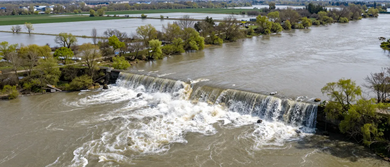 Aerial view of a swollen river overflowing, cascading over a weir into flooded surrounding spring fields