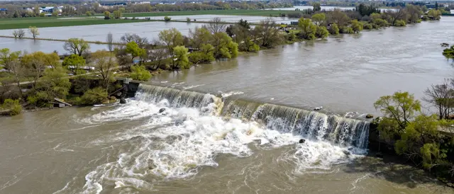 Aerial view of a swollen river overflowing, cascading over a weir into flooded surrounding spring fields