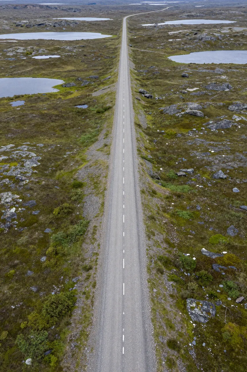 A straight long gravel road stretches through rocky green tundra dotted with small shallow lakes.