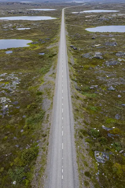 A straight long gravel road stretches through rocky green tundra dotted with small shallow lakes.
