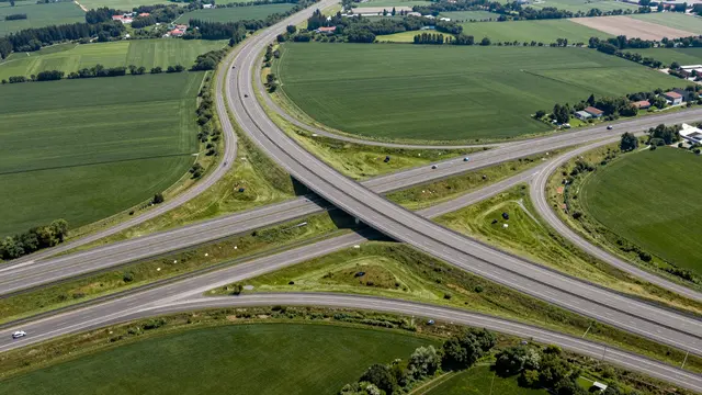 Aerial top-down view of a multi-lane highway interchange winding through green rural farm fields on a sunny day.