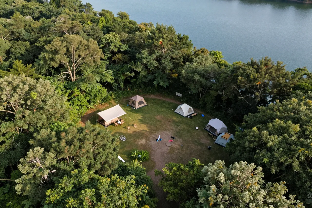 A high-angle aerial view of a grassy forest clearing with tents beside a calm blue lake surrounded by dense green trees.