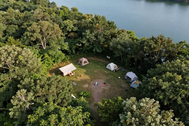 A high-angle aerial view of a grassy forest clearing with tents beside a calm blue lake surrounded by dense green trees.