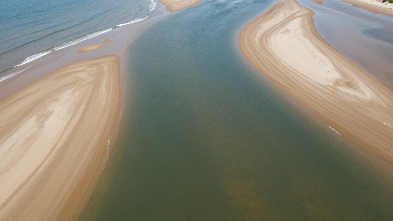 Aerial view of a river mouth where calm greenish water meets the blue ocean between curved sandy sandbars