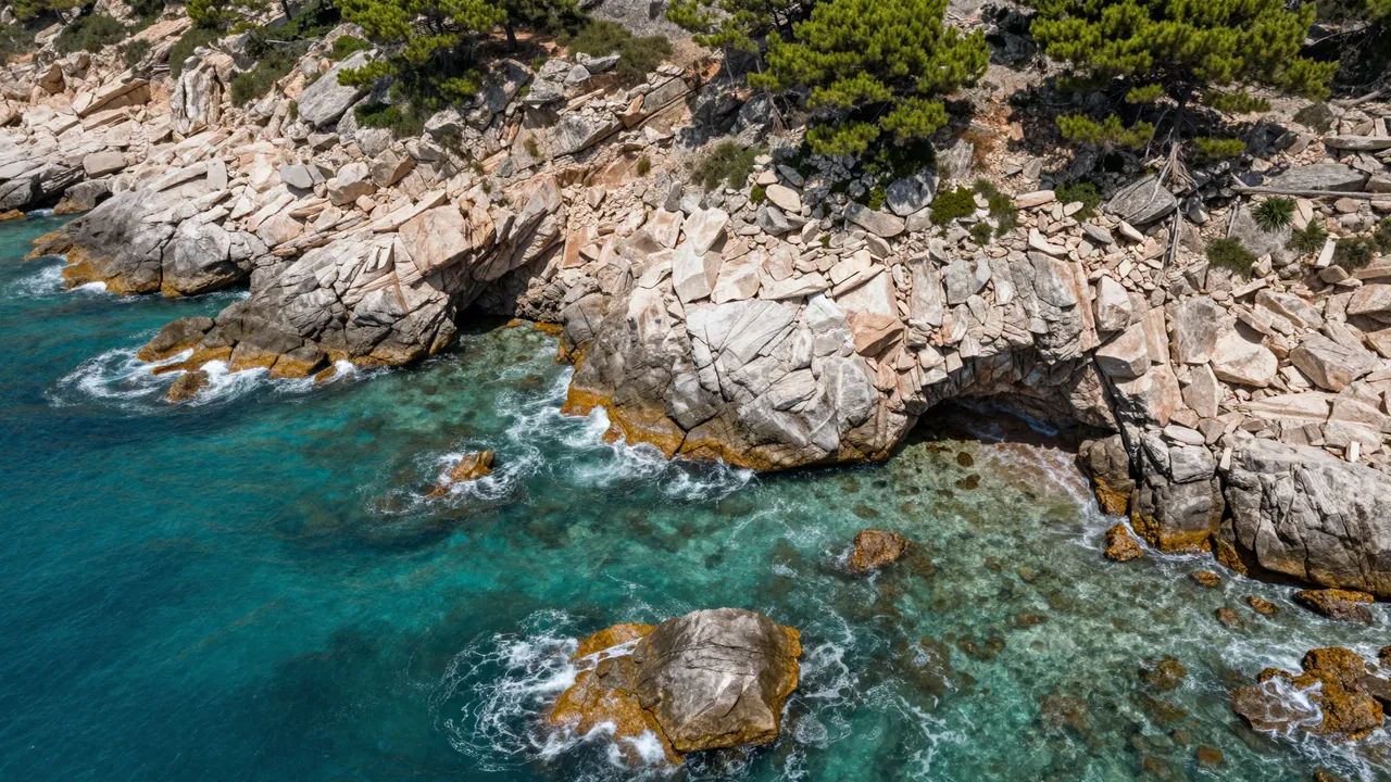 Aerial view of turquoise coastal coves framed by pine-topped rocky cliffs with breaking surf