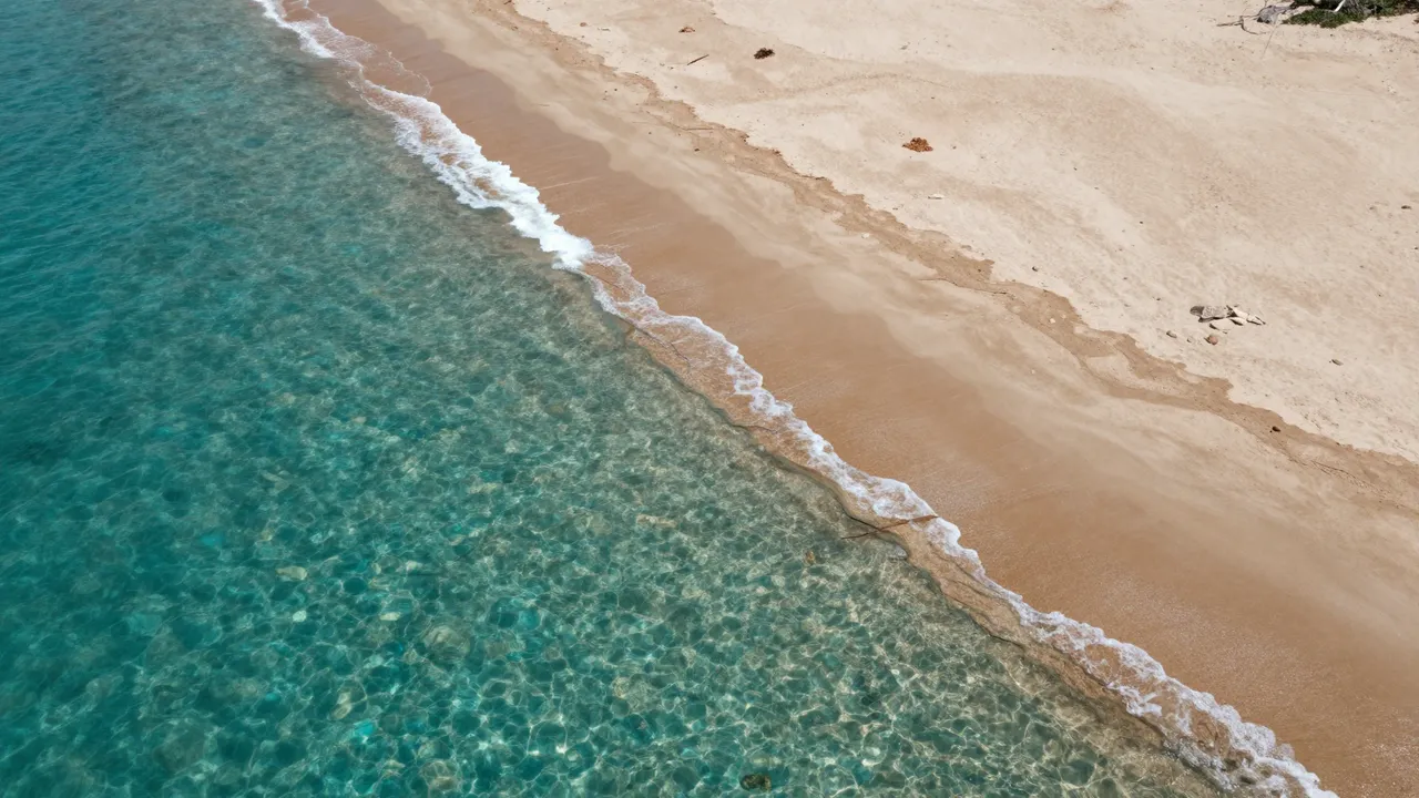 Aerial view of gentle white surf lapping a sandy beach next to clear turquoise shallow water