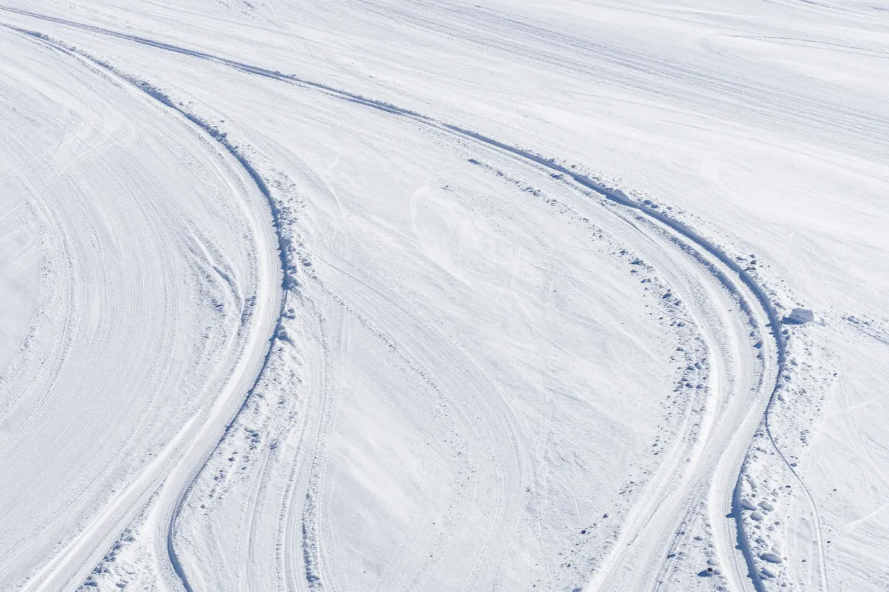 Curved fresh ski tracks wind across a wide empty sunlit snowfield viewed from above.