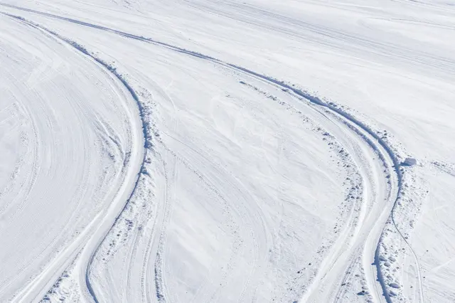 Curved fresh ski tracks wind across a wide empty sunlit snowfield viewed from above.