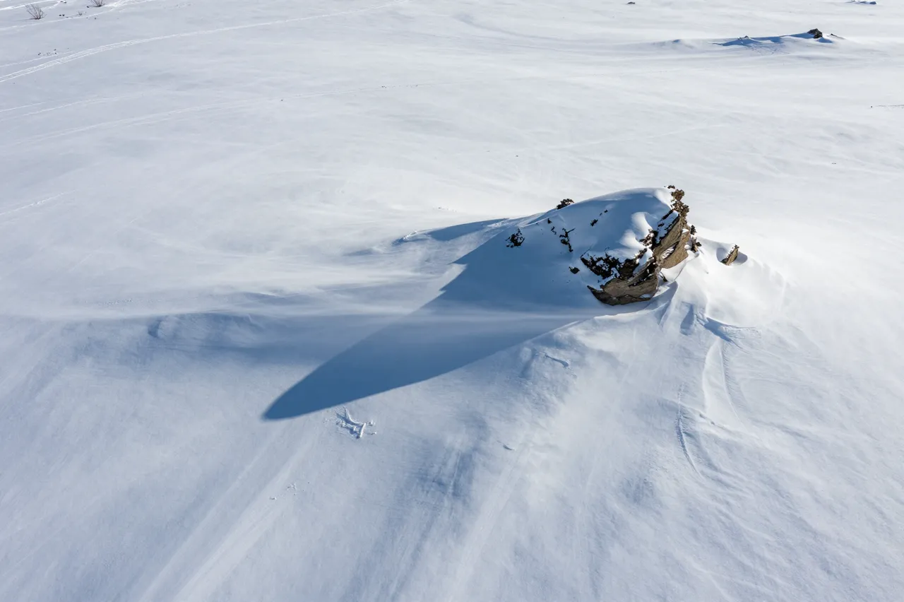 Aerial view of wind-sculpted snow drifts surrounding a lone rocky outcrop on a bright frozen plateau