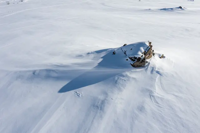 Aerial view of wind-sculpted snow drifts surrounding a lone rocky outcrop on a bright frozen plateau