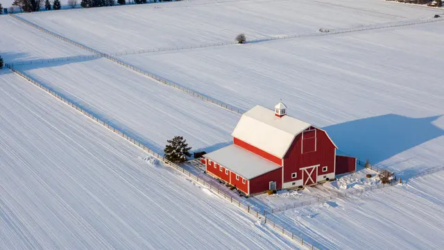 Aerial view of a red barn sitting in snow-covered farm fields at golden dawn with fenced property lines.