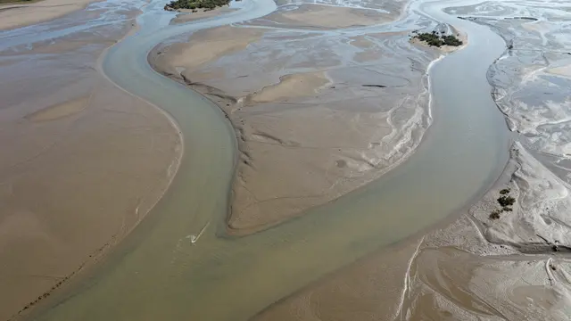 A high-angle aerial view shows winding shallow water channels across wide muddy tidal flats with small vegetated islets.