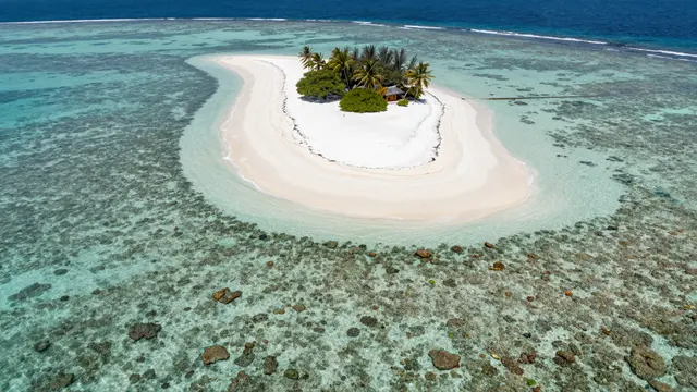 A high angle aerial view of a small tropical island with white sand, palm trees, and surrounding turquoise coral reef.