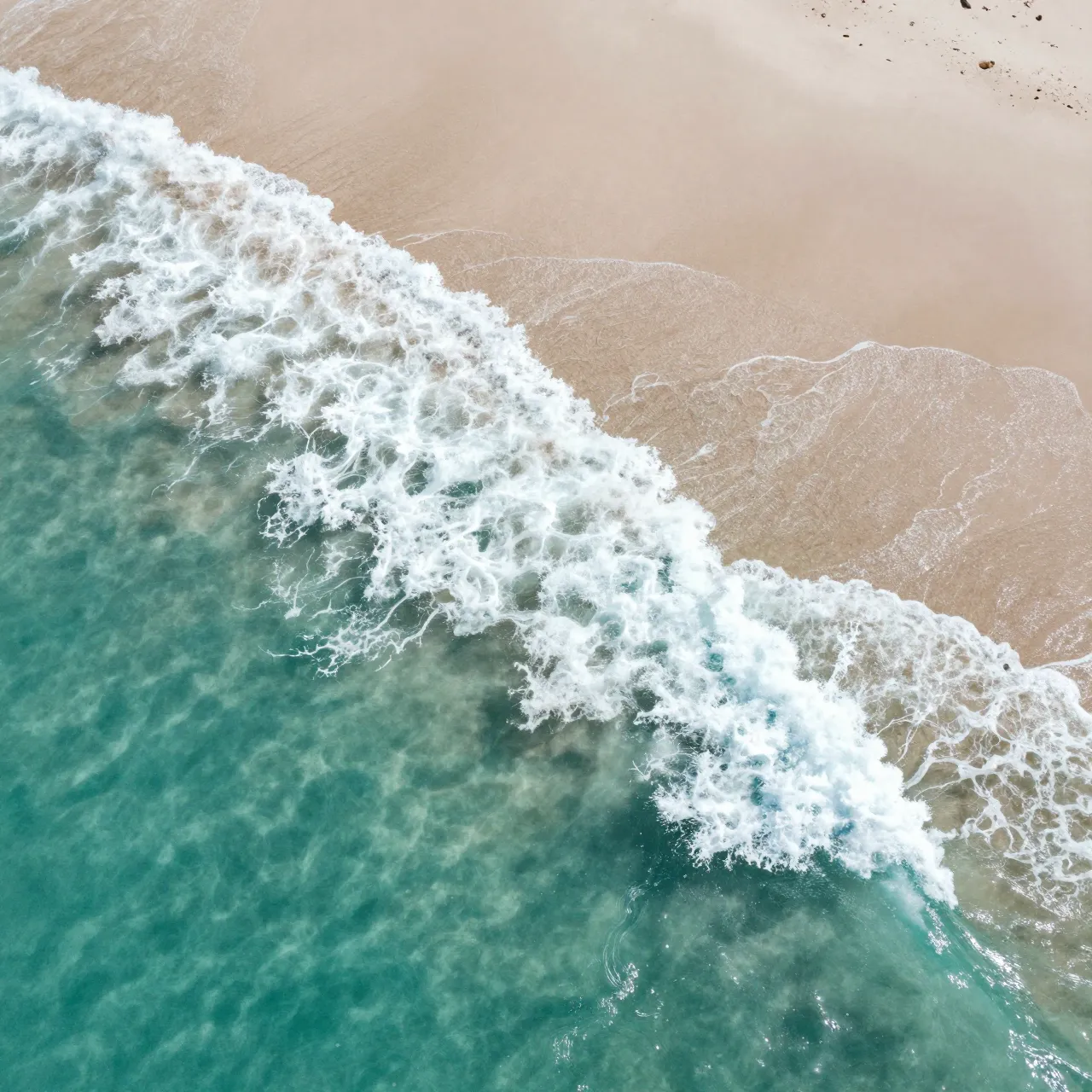 Top-down aerial view of foamy turquoise ocean waves rolling onto pale sandy shore