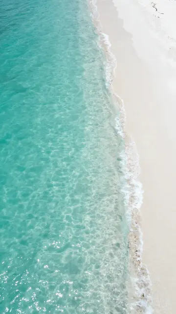 Top-down vertical view of clear turquoise ocean gently lapping against a bright white sandy beach.