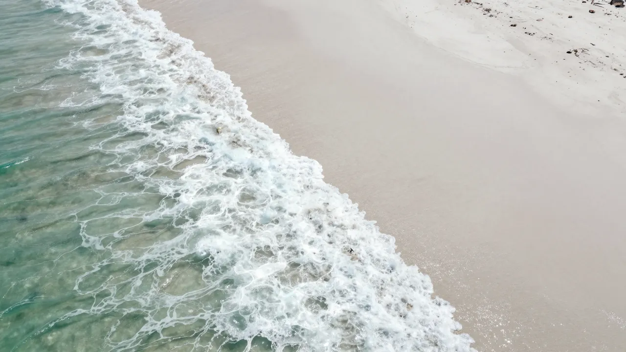 Aerial perspective of foamy turquoise ocean waves meeting a pale curved white sand beach