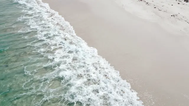 Aerial perspective of foamy turquoise ocean waves meeting a pale curved white sand beach