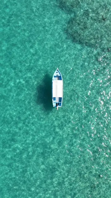 A small light blue and white boat floats centered in crystal clear turquoise lagoon water viewed from directly above